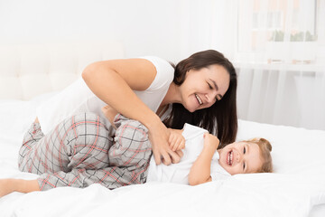 adult mother is tickling her little active daughter in bed at home, having fun, leisure activity...