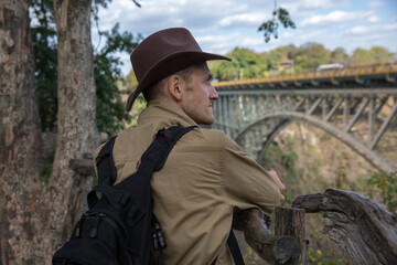 a man tourist in a cowboy hat and with a backpack stands with his back to us and looks at the bridge
