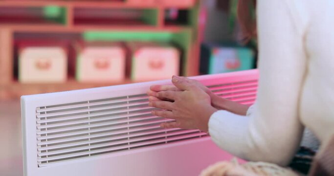 Young woman warming hands near electric heater at home