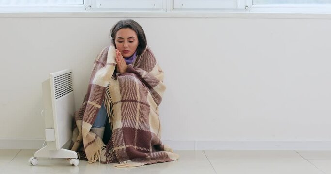 Young woman wrapped in warm plaid sitting near switched on electric heater in room