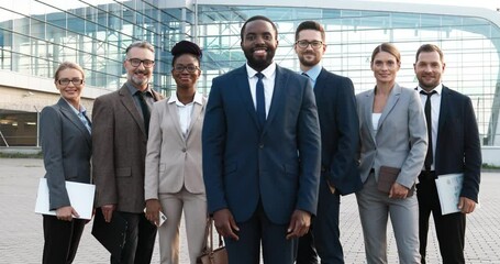 Portrait of successful African American male young boss with his team of males and females company workers. Mixed-races businessmen and businesswomen smiling to camera outdoor at business center.