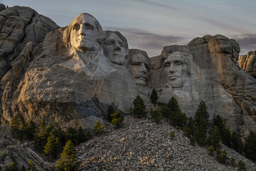 A view of the Mount Rushmore National Memorial in South Dakota during sunset. The President's faces are illuminated by the sun and the sky has dark clouds and hues.