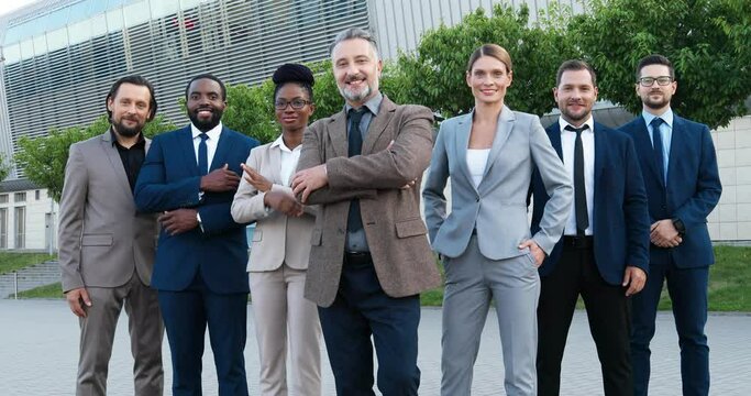 Portrait of successful rich gray-haired Caucasian male boss with team of multi ethnic males and females company coworkers. Mixed-races businessmen and businesswomen smiling to camera outdoor.