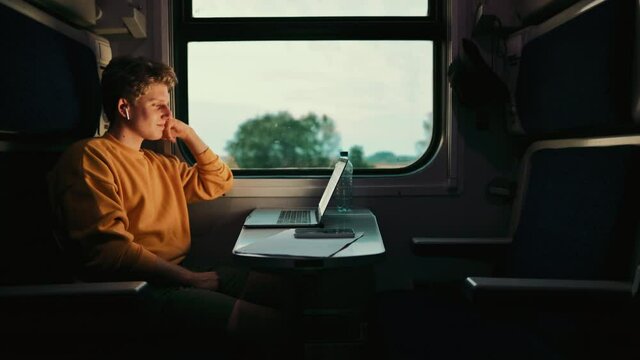 positive young man in a yellow sweatshirt rides in a train compartment and watches funny content on a laptop with a smile on his face, listens in wireless headphones.
