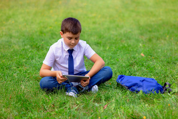 A happy schoolboy in a white shirt sits on green grass and play a tablet