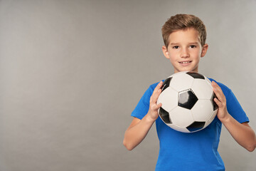 Adorable boy with soccer ball standing against gray background