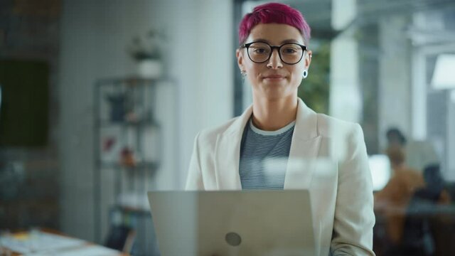 Modern Office: Portrait of Beautiful Authentic Specialist with Short Pink Hair Standing, Holding Laptop Computer, Looking at Camera, Smiling Charmingly. Working on Design, Data Analysis,Plan Strategy 