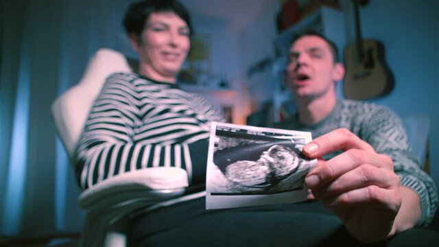 A pregnant woman is sitting in a chair, next to her husband is holding an ultrasound photograph of a child. Show the sympathy of young parents. Continuation of the human race. Young couple posing.