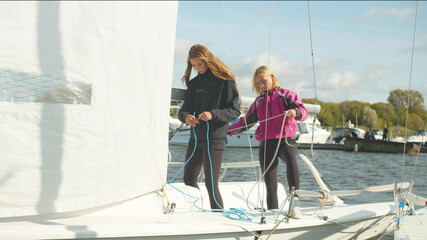 Obraz premium Two cute girls stand on the stern of a white sports yacht and pull the ropes to control the sail