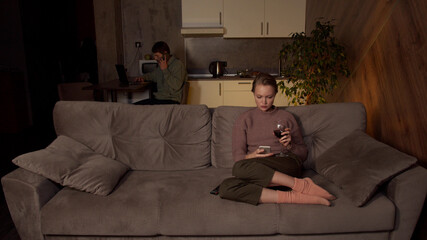 A girl in cozy clothes is sitting on the sofa in the living room, drinking wine and watching TV. Her husband is at the kitchen table, working on a laptop, making work calls on a smartphone.
