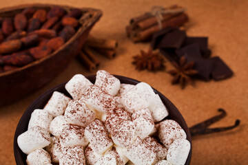 Cup of cacao drink with white marshmallows, chocolate, spices, cocoa pod and cacao beans on a cork background.
