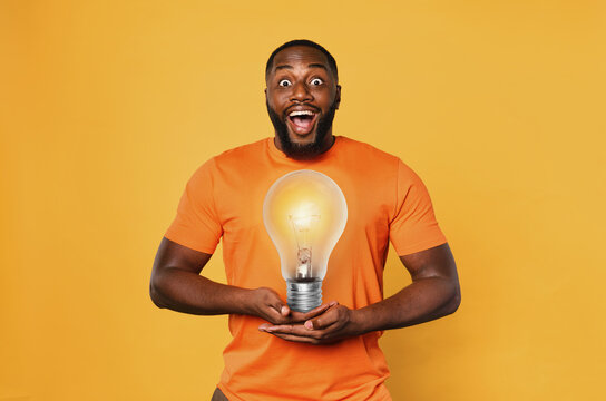 Happy afro man holds a lightbulb with hands. Concept of idea and creativity. Orange background