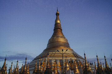 a large ornate building with a gold roof and a blue sky with Shwedagon Pagoda in the background