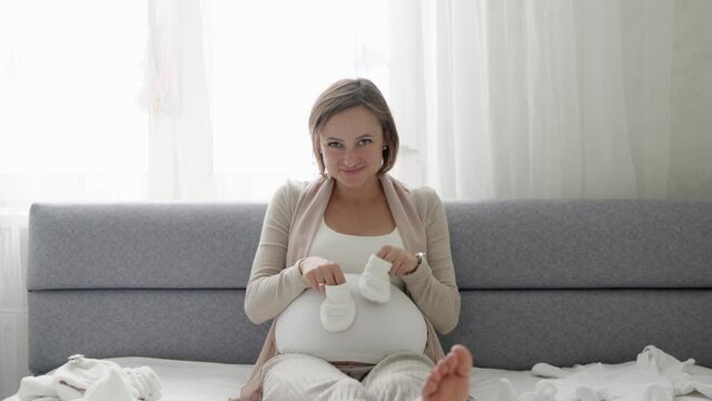 YounYoung pregnant woman with small baby shoes sitting on sofa at home. Expectant mother is preparing for childbirth. High quality 4k footage