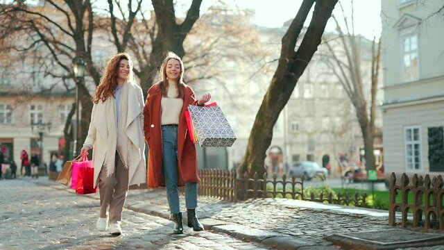 Two happy young women walking around the city after shopping on a city street in coats with gift bags in cold autumn winter or spring weather 2 cheerful girls go outside from a sale in a mall or store