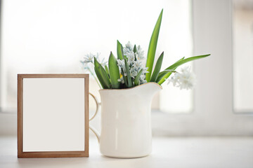 White ceramic jug with Fresh blue spring flowers Scilla siberica on windowsill on background of window. wooden empty frame with white background. Mock up