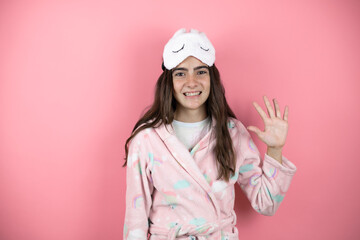 Pretty girl wearing pajamas and sleep mask over pink background showing and pointing up with fingers number five while smiling confident and happy