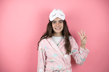 Pretty girl wearing pajamas and sleep mask over pink background showing and pointing up with fingers number three while smiling confident and happy