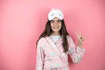 Pretty girl wearing pajamas and sleep mask over pink background showing and pointing up with fingers number two while smiling confident and happy