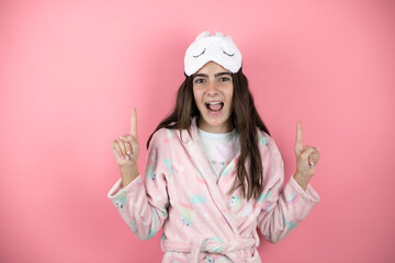 Pretty girl wearing pajamas and sleep mask over pink background amazed and surprised looking at the camera and pointing up with fingers and raised arms