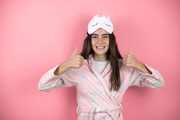 Pretty girl wearing pajamas and sleep mask over pink background smiling confident doing the ok signal with her thumbs