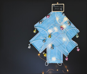 View of a Christmas tree made of blue surgical masks decorated with colorful Christmas baubles and Christmas lights against black chalkboar background. Christmas during pandemic or medical background.