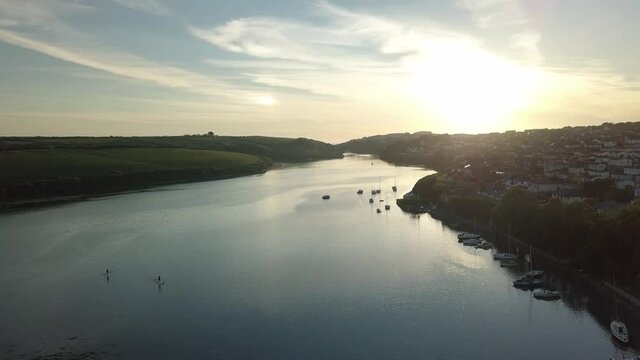 Two People Paddleboarding At River Gannel With Boats Moored On Riverside Near Newquay Town On Sunrise. - aerial