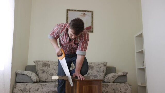 Carpenter cutting a wooden plank with hand saw at home in the living room. Video. Confident young handyman using saw during repair of apartments.