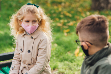 Little boy and girl in in protective masks looking at each other while resting in park on autumn...
