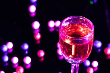 Crystal Wine Glass Goblet Full Of Red Color Alcohol Shot For Late Night Christmas Happy New Year Party Theme With Reflection Of Colorful Decoration In Blur And Vibrant Light On Dark Black Background