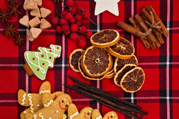 Christmas greeting card. Gingerbread men, hearts made of brown sugar, fir shaped cookie, dried orange slices, red berries, vanilla beans, cinnamon sticks on a red fabric tartan background. Top view.