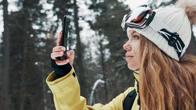 A girl in a white hat and a snowboard mask in the forest photographs nature on the phone in winter
