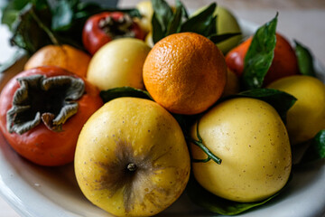 Brightly colored, orange and yellow, winter fruit: persimmon, apples, lemons and oranges arranged on a platter. Some green leaves. 