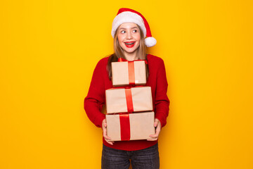 Pretty overjoyed woman holding giftbox close to chest wear knitted jumper and santa hat isolated yellow background