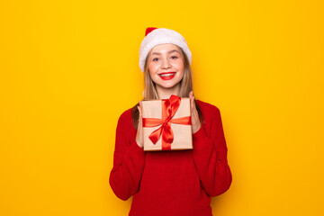 Pretty overjoyed woman holding giftbox close to chest wear knitted jumper and santa hat isolated yellow background
