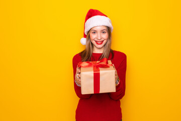 Pretty overjoyed woman holding giftbox close to chest wear knitted jumper and santa hat isolated yellow background