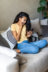 Excited woman reading text on a phone lying on a couch in the living room at home