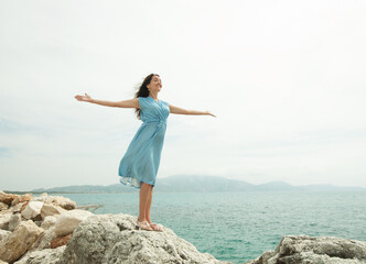 A young woman with dark hair in a blue dress stands on a stone with her arms outstretched, next to the sea