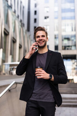 Attractive young businessman wearing a suit sitting on a bench outdoors at the city street, talking on mobile phone while drinking takeaway coffee
