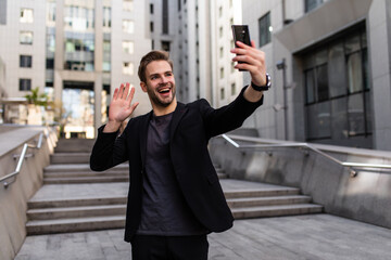 A successful handsome businessman in a suit, holding a mobile phone in his hand, making a selfie, a background of skyscrapers. Concept: communication, contacts, internet, chat, network.