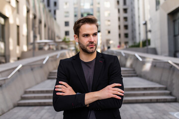 Caucasian male banker with crossed hands standing at urbanity in financial district and thoughtful looking away, pensive businessman thinking about corporate lifestyle pondering in downtown