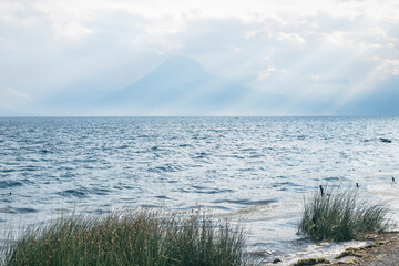 View from the grass coast over lake Atitlan to the volcano with sunrays at Panajachel, Guatemala