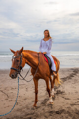 Beautiful caucasian woman riding horse on the beach. Female wearing white clothes. Copy space. Sunset time on the beach. Outdoor activities.