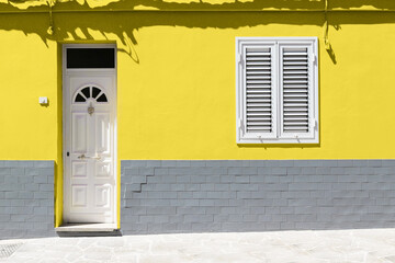 White wooden front door and white window on yellow wall with gray brick. Color trend 2021.