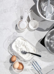 Top view of bakery equipment for a homemade healthy dessert. Ingredients prepared on a white and clean marble countertop. Eggs, wheat flour, hand mixer, spatula, mixing bowl, measuring cups, and scale