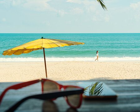 Yellow beach umbrella against the background of the beach, sea and a walking woman. In the foreground are glasses and a smartphone at the bar desk - Powered by Adobe