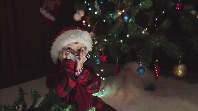 coronavirus, covid-19 in the new year. a little girl in a red Santa Hat and mask is sitting under the Christmas tree at home.