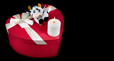 Obraz na płótnie Canvas Red heart shaped gift box, candle burning and toy cow bull Isolated on black background. Valentine day in 2021. Copyspace for text