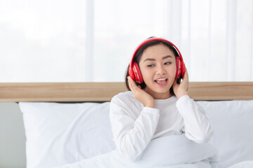 Happy young female teenage girl in headphones listening to music from smartphone on bed at home. Young beautiful woman with headphones relaxing in her bed, listening to music with smart phone.