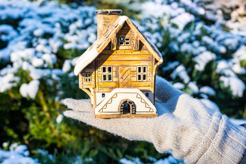 A toy ceramic house on a female palm in a warm mitten against the background of a blurred evergreen thuja in the snow